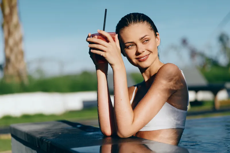 Young woman in a white sports bra relaxes at the edge of an outdoor pool holding a drink with a straw, enjoying a sunny day with a calm and content expression.