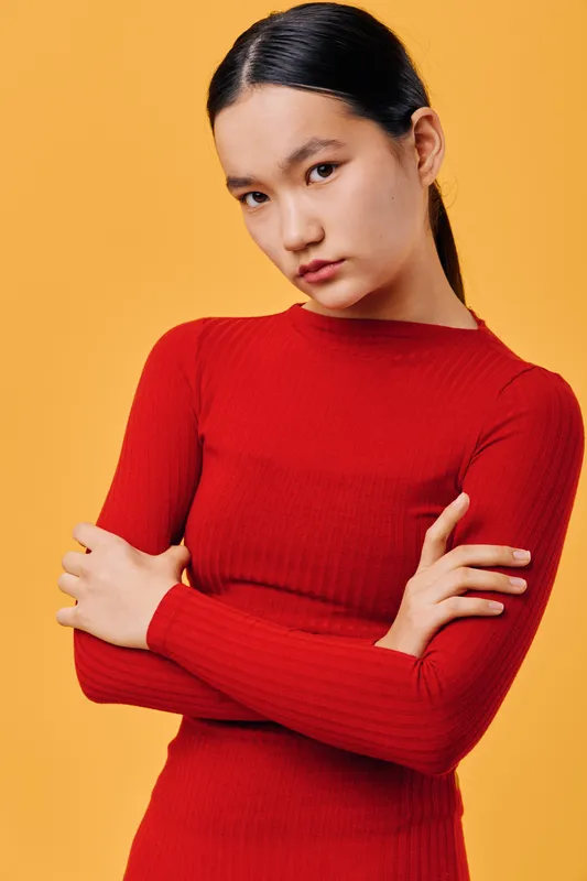 Portrait of young woman with straight black hair in red sweater crossing arms and looking serious on solid yellow background. Asian female model posing with natural expression in studio light.