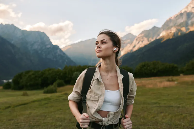 Young woman on a hike through mountains, wearing casual outdoors attire and backpack, gazing at distant peaks, capturing a fresh nature driven lifestyle moment in the open air and light
