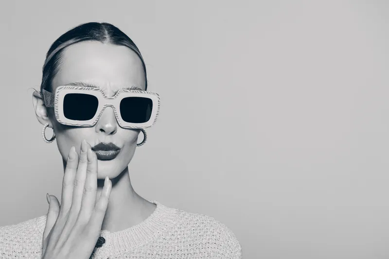 Stylish young white woman with pale skin wearing large square sunglasses and hoop earrings, dressed in knitted sweater, posing with hand near mouth in studio.