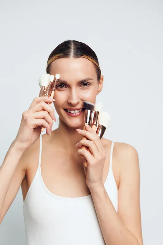 Young woman with blonde hair smiles while holding several makeup brushes near her face against a plain white background in a bright studio setting.