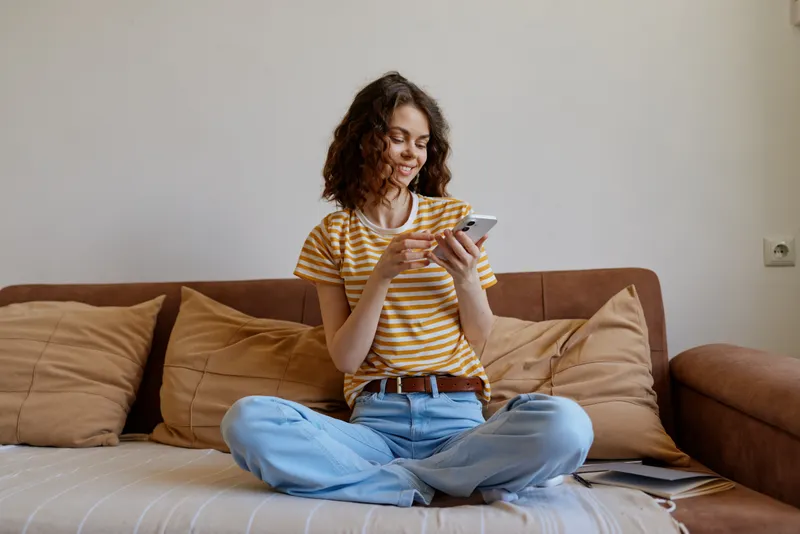 Young woman sits cross legged on a living room couch at home, wearing a striped tshirt and jeans, smiling while checking her smartphone in a cozy casual moment.