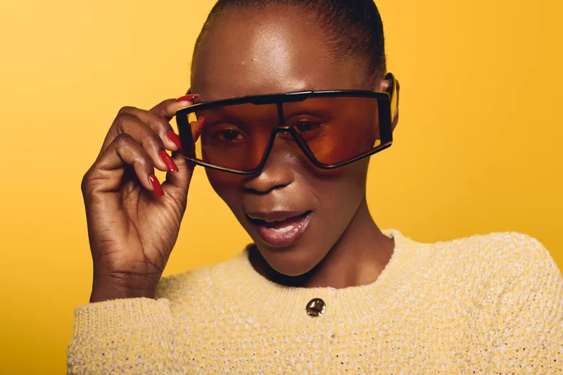 Stylish black woman with dark skin wearing oversized orange sunglasses and yellow sweater poses confidently against yellow background. Concept of fashion, beauty, and modern style.