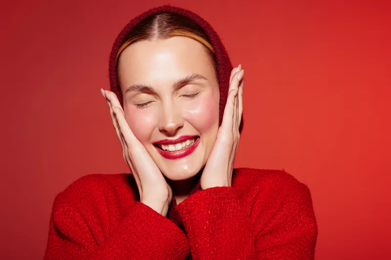 Smiling woman in red sweater with closed eyes touches face with hands on red background. Concept of happiness, beauty, skincare, and cozy winter fashion with warm colors and soft lighting.