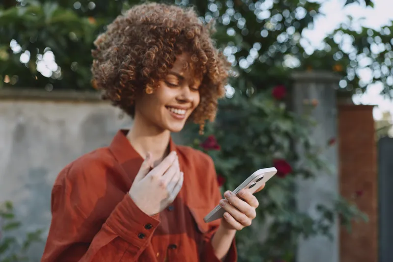A cheerful woman in a casual red shirt stands outdoors in a garden, checking her phone with a bright smile. Candid lifestyle moment for everyday connectivity