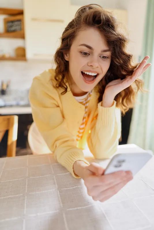 A cheerful woman in a cozy home setting holds a smartphone, smiling at the screen, capturing a candid selfie. Bright natural light enhances a relaxed, everyday lifestyle moment.