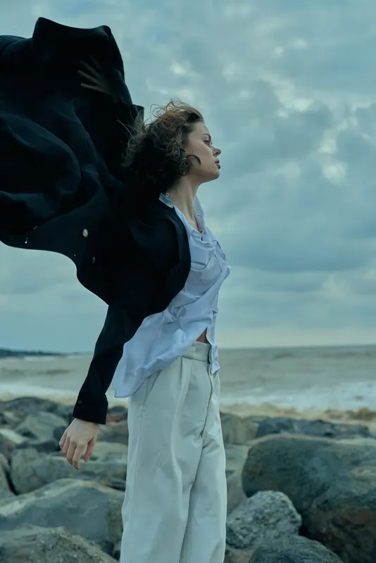 Woman standing on rocky shore with coat flowing in wind, white blouse and light pants, ocean backdrop, motion and freedom, fashion moment in dramatic outdoors