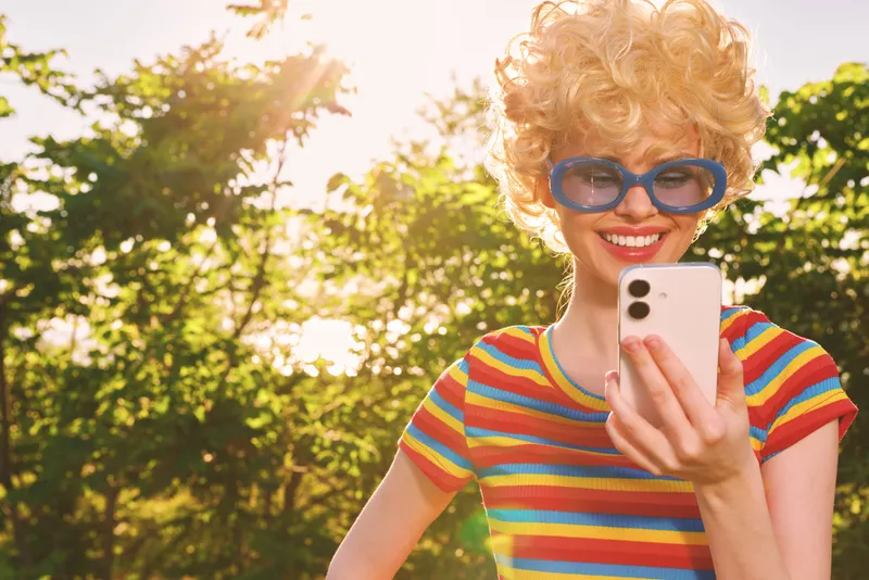 Woman outdoors in a striped shirt wears retro goggles and selfies in a sunny park, capturing a cheerful lifestyle moment with warm film color and bright natural light. Soft glow.