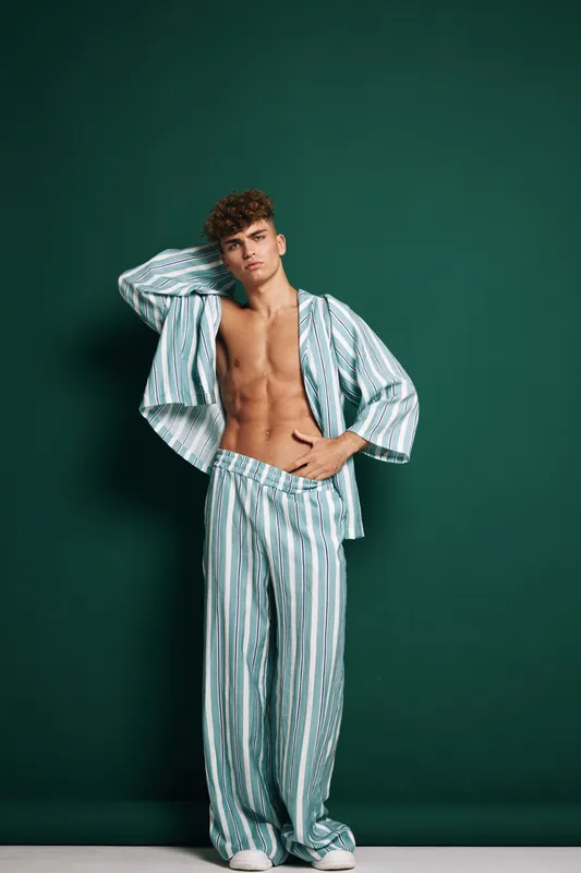 Young man posing in striped pajamas with bare chest and relaxed posture against green background. Casual sleepwear with loose pants and shirt with arms back and hand on abdomen.
