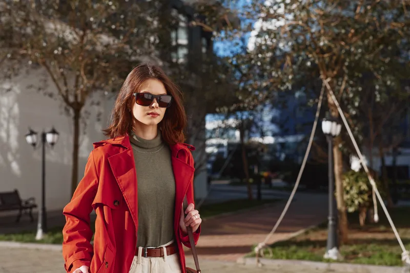 Vibrant woman outdoors in a red coat and sunglasses, walking through an urban park, autumn light, confident pose, street fashion moment with soft breeze and natural colors