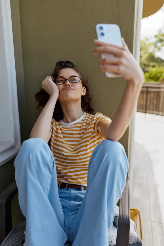 A casual woman sits on a balcony taking a selfie with a smartphone, wearing a striped shirt and light blue jeans. Concept: relaxed lifestyle and home daily life.