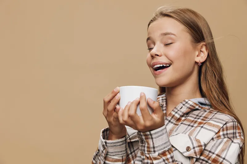 Girl holding white cup with eyes closed enjoying aroma or taste of warm coffee or tea in cozy plaid shirt against beige background. Relaxation and comfort moment indoors.