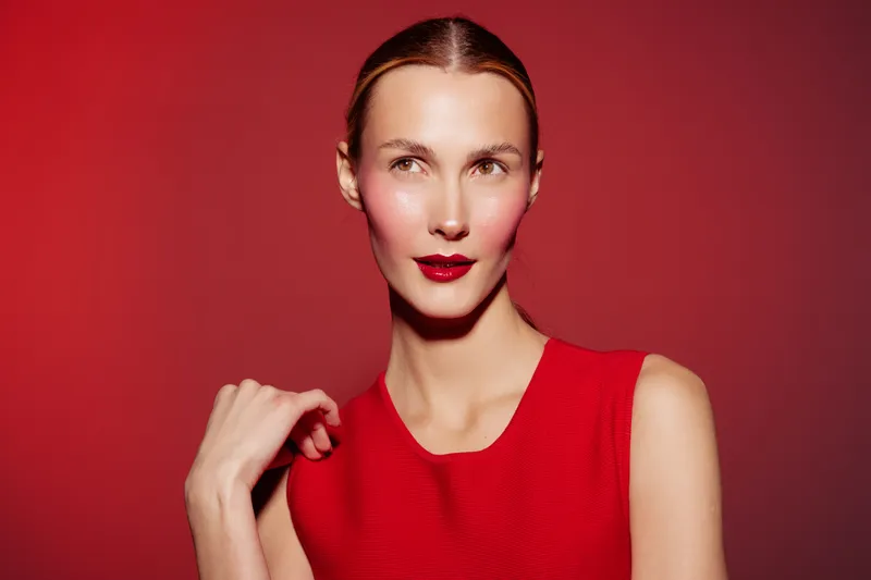 Portrait of a confident woman in red dress with red lipstick on red background. She looks away with a slight smile, showcasing elegance and fashion style in studio lighting.