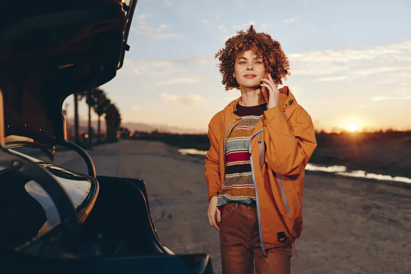 Woman lifestyle outdoors near car at sunset, curly hair, casual jacket and denim, standing by the open trunk on a road, warm golden hour light, peaceful travel mood at dusk