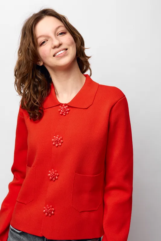 Smiling woman with wavy brown hair wearing a bright red sweater with decorative buttons stands against a white background. Concept of casual fashion and cheerful mood.