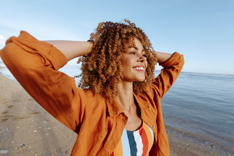 A happy young woman with curly hair wearing an orange shirt enjoys a sunny day at the beach with a clear blue sky and calm sea in the background.