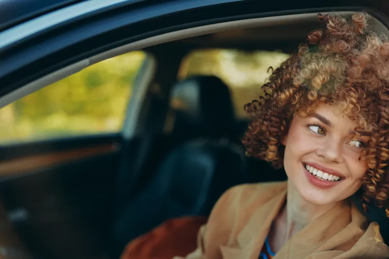 Bright woman in a car, smiling with curly hair, sunlit interior moment, relaxed pose, warm colors, soft light, candid lifestyle scene capturing joy, style and optimism retro film color warm ambience