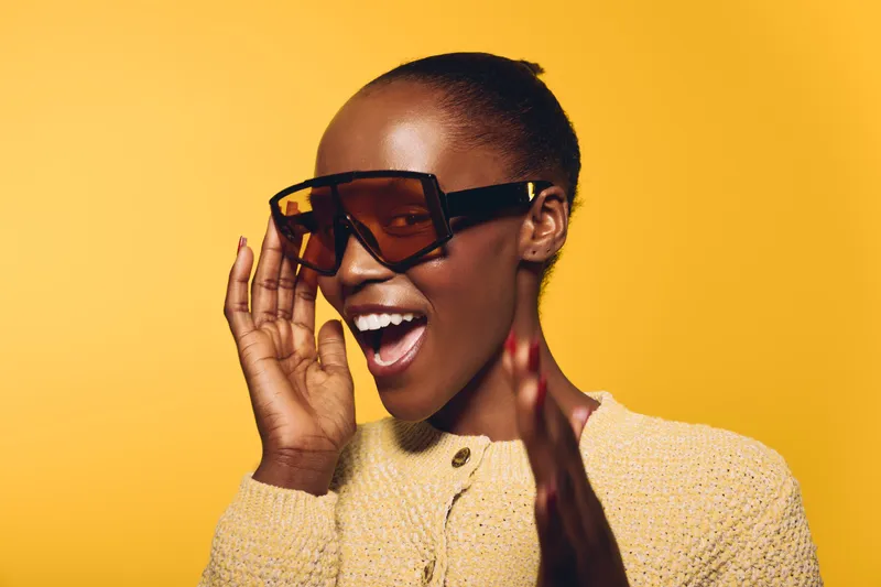 Happy African woman with dark skin wearing large sunglasses and a beige sweater smiles joyfully on a yellow background. Concept of fashion, style, and positive emotions.