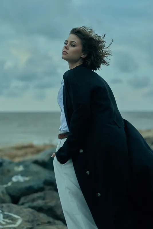 Windy coastal scene with a stylish woman standing on rocks, coat billowing, sea in the background, moody sky and fashion mood