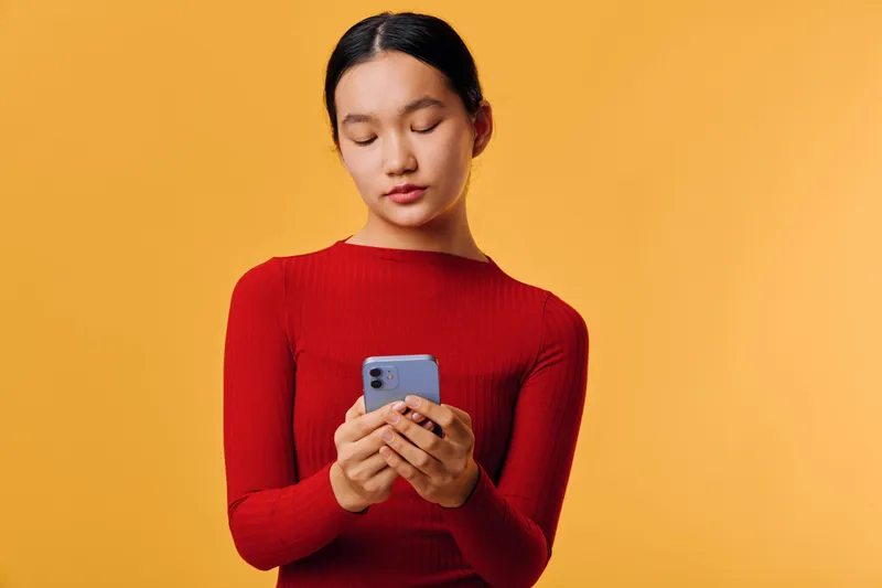 Young woman in red sweater holding smartphone looking down with concentration against orange background for communication, technology and social media concept.