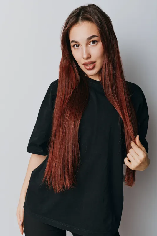 Young woman with long straight red hair wearing black oversized t shirt posing in studio with gray background. Casual style, beauty, fashion and confident model portrait.