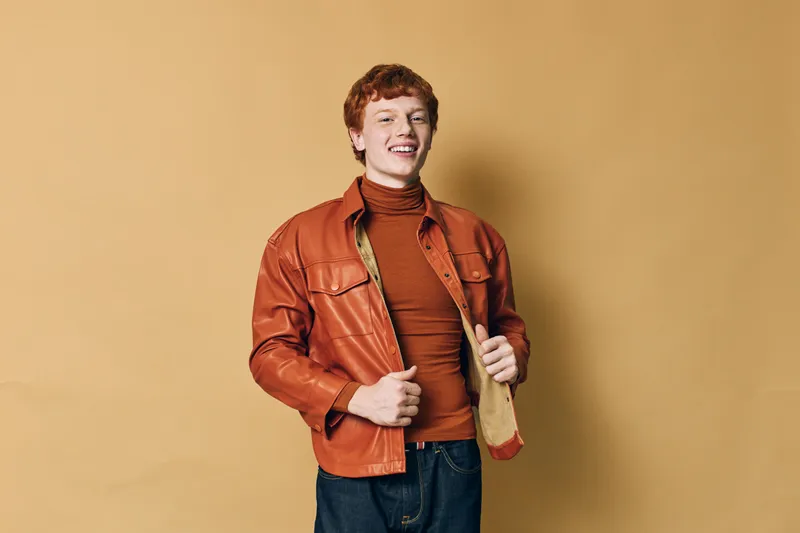 Young man with red hair wearing orange leather jacket and turtleneck smiling confidently standing against beige background in casual stylish outfit for fashion portrait.