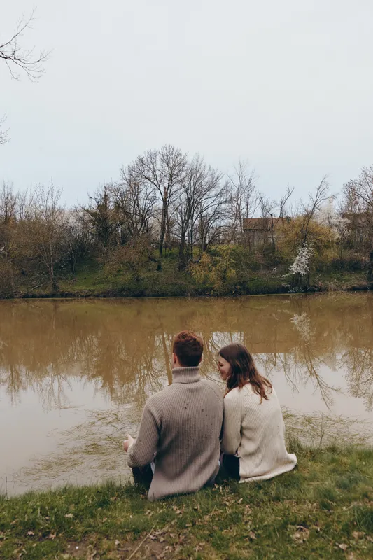 Couple sitting by a calm lake in autumn, back view of lovers in cozy sweaters, reflecting a peaceful park scene with trees and water, warm intimate moment, lifestyle romance and serenity.