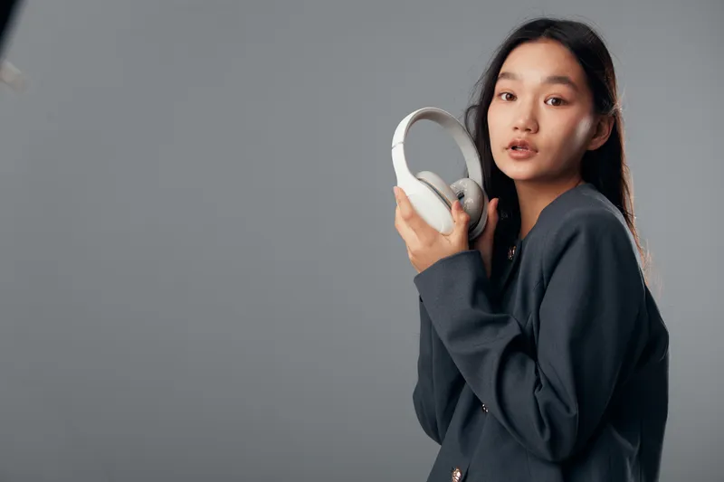 Young woman holding white wireless headphones indoors, wearing dark outfit, looking at camera with modern technology and casual fashion style on gray background.