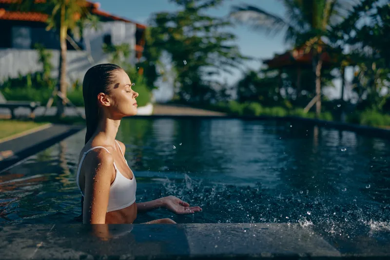 Woman relaxes in a sunlit pool, standing waist high, calm expression, wearing a white swimsuit, tropical backdrop with palm trees, gentle splashes around, tranquil lifestyle moment ideal for leisure,