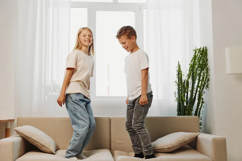 Two happy children jumping on sofa in bright living room with natural light and green plant. Playful kids enjoying leisure time indoors at home.
