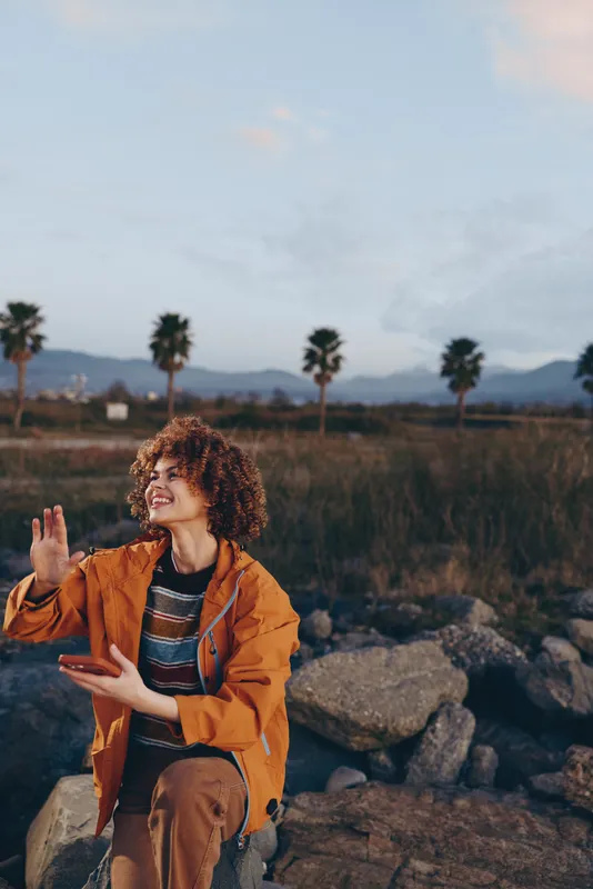 Woman in orange jacket with curly hair stands outdoors on rocky ground at sunset, smiling and waving hands, travel lifestyle. Palm trees, dry grass, and distant hills frame the calm nature scene
