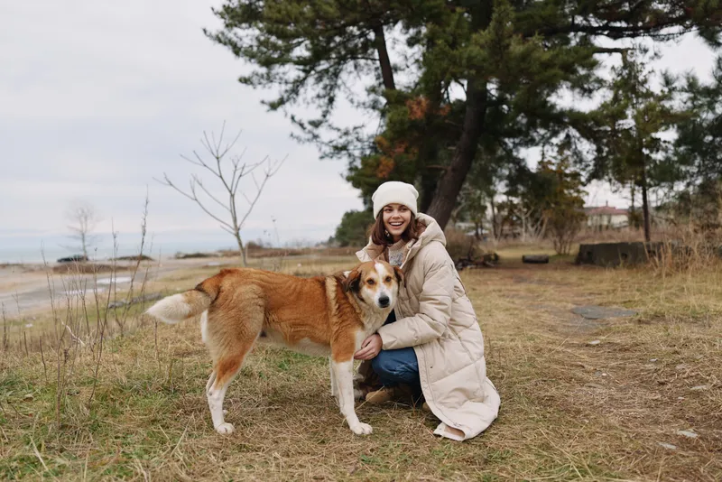 Woman in a warm coat and hat kneels with a friendly dog in an open park by the coast, enjoying nature and a quiet outdoor moment together