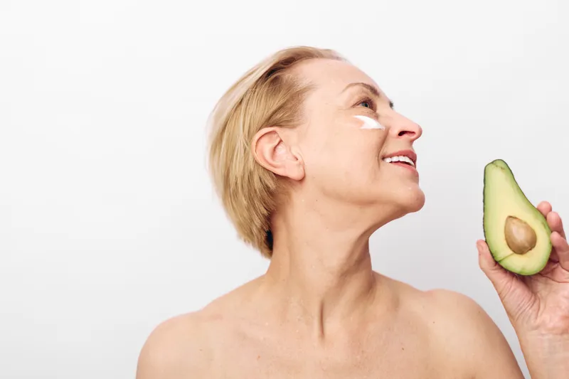 Smiling mature woman applies natural face cream holding avocado half close to skin, isolated on white. Concept of organic skincare, healthy lifestyle, and natural beauty with fresh fruit ingredients.