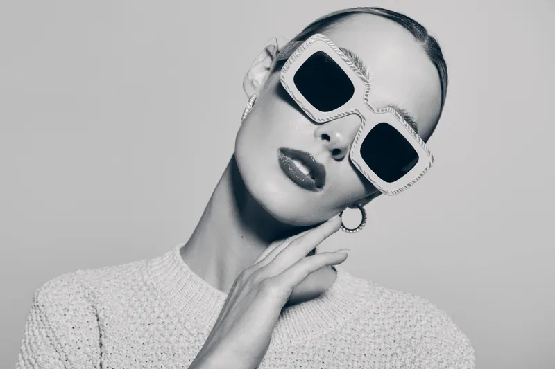 Stylish white woman with pale skin wearing large sunglasses and textured sweater poses with hand near face in monochrome studio portrait. Fashion and elegance concept.