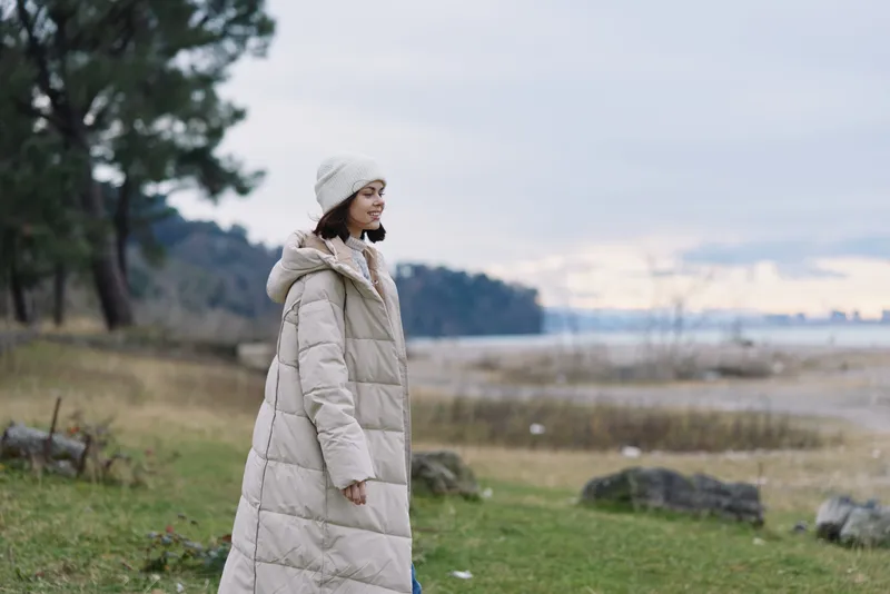 Woman in a long beige puffer coat stands along a windy seaside park, looking toward the horizon, capturing a calm landscape, winter mood and outdoor fashion portrait