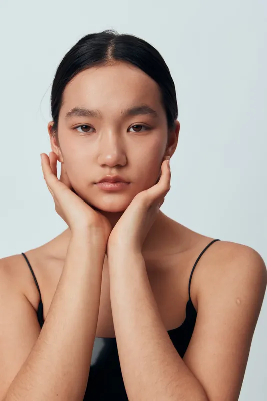Young woman with natural skin and dark hair posing with hands on face for beauty portrait and skincare concept against neutral background in studio light.
