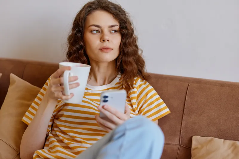 Young woman with curly hair sits on a cozy sofa, wearing a striped yellow shirt, holding a white mug while scrolling on her smartphone in a bright living room.