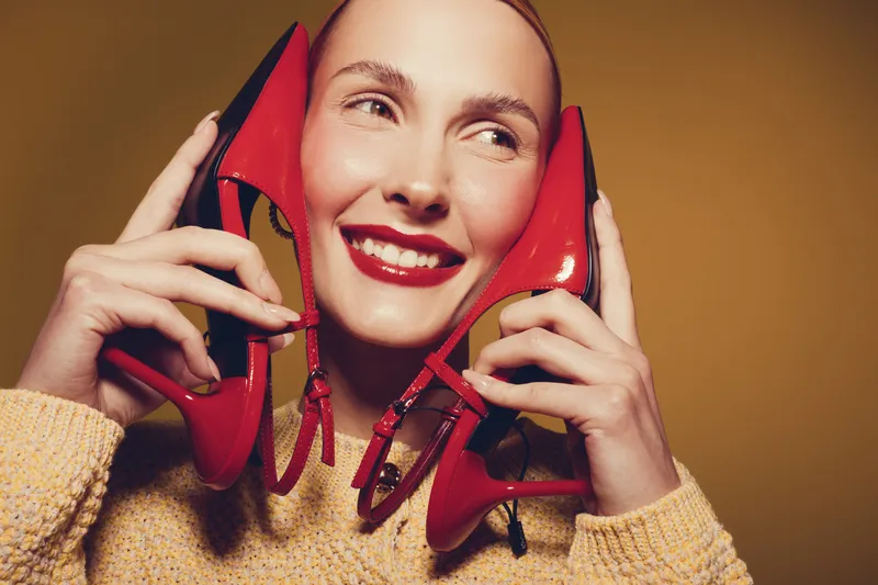 Smiling woman holds red high heel shoes near face, wearing yellow sweater. Close-up portrait on brown background. Concept of fashion, style, and joyful emotions.