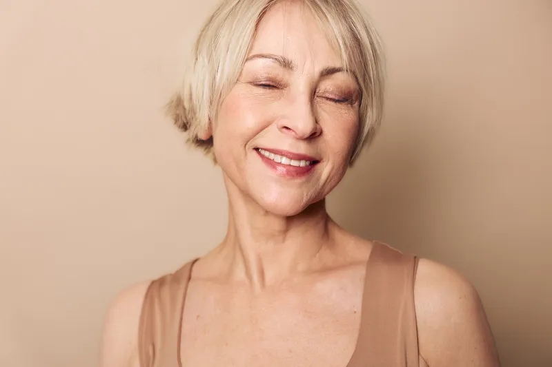 Smiling mature woman with closed eyes enjoys calm moment. Soft beige background highlights natural beauty, skin glow, and peaceful expression. Concept for wellness and self-care.