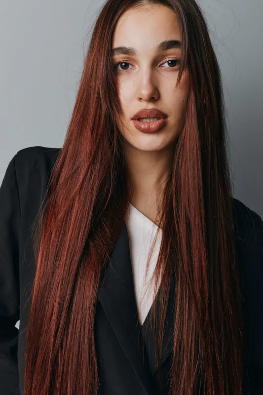 Woman with long straight hair wearing black blazer and white top posing in studio with neutral background. Portrait of young female with natural makeup and confident expression.