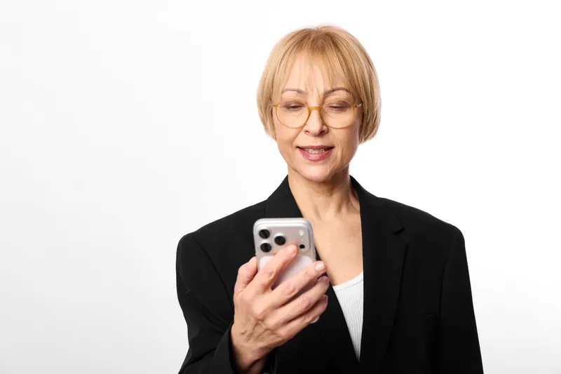 Woman elderly smartphone glasses portrait business suit communication technology smiling, holding smartphone in studio, white background, professional corporate portrait.