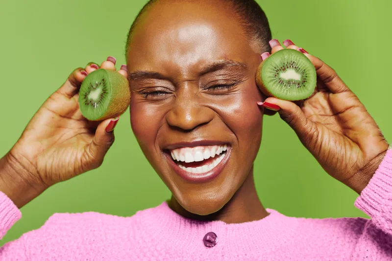 Happy dark skinned African woman with short hair smiling widely holding two halves of kiwi fruit near her face on green background. Concept of healthy lifestyle, fruit, and joy.