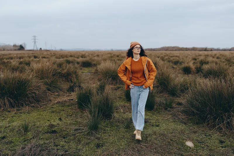 woman outdoors walking lifestyle in autumn field, grass path and greenery, wearing orange jacket and sweater, relaxed confident smile, fullbody portrait on meadow trail, daylight clouds