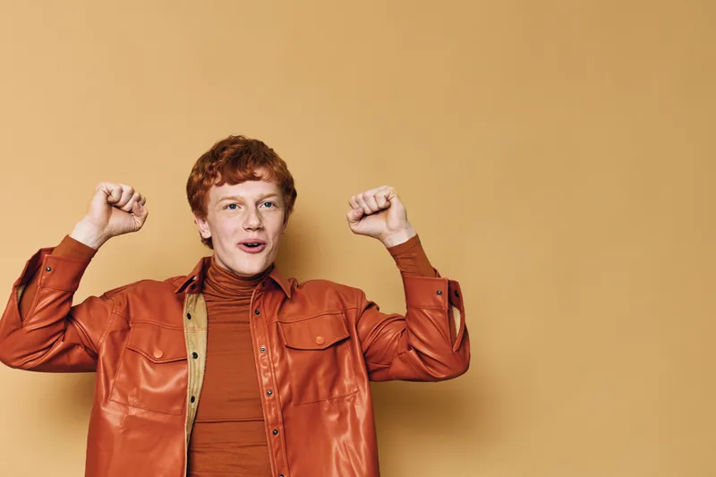 Young man with red hair wearing orange shirt and brown turtleneck sweater raising fists with excited face on beige background smiling and celebrating success.