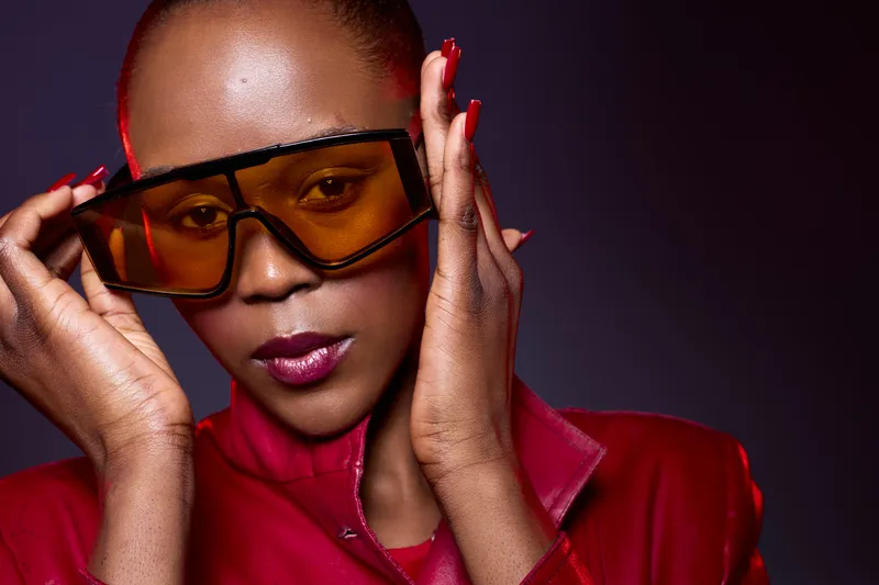 Stylish dark skinned African woman wearing large orange sunglasses and red leather jacket poses confidently in studio. Fashion portrait with glossy lips and manicured nails on dark background.