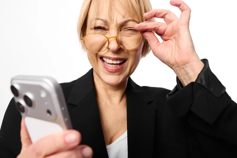 Mature woman adjusting glasses, smiling and holding a smartphone for video calls. Close up portrait on a white studio background. Expression, communication, and technology.