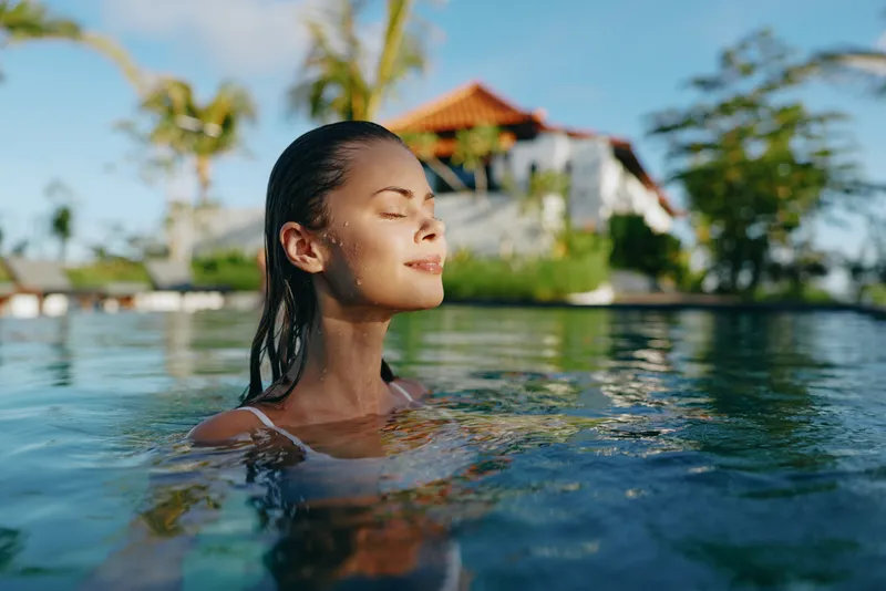 Calm woman swims in a sunny pool, eyes closed, enjoying warm water and gentle reflections on blue tiles, embodying wellness, relaxation, and carefree summer vibes today.