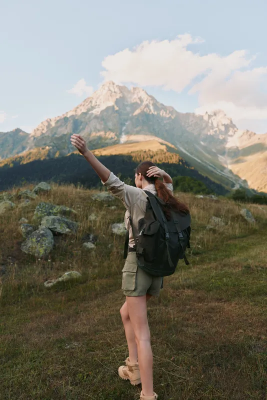 A woman with backpack hikes through alpine meadow, arms raised, rugged mountains in the background under a bright sky, conveying adventure and outdoor lifestyle.