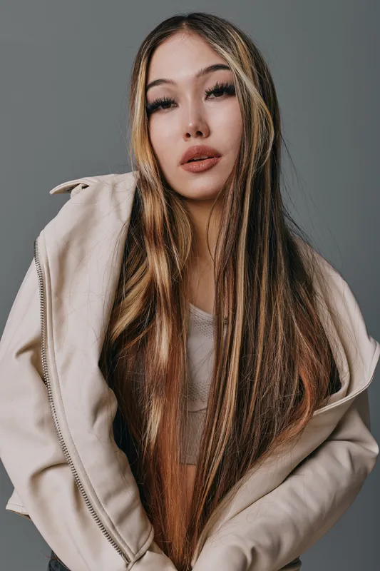 Woman with long straight hair wearing beige jacket and top posing confidently against gray background with casual style and natural makeup for fashion or portrait photography.