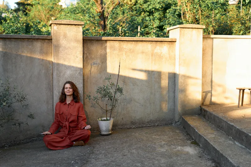 Woman sits cross legged in a red dress beside a concrete wall, calm street style scene, minimalist outdoor mood, casual solo portrait, natural light, serene atmosphere